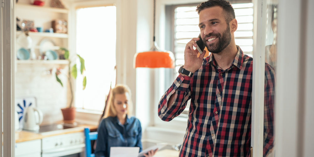 Handsome,Man,Talking,On,The,Phone,At,Home. Handsome man smiling while talking on the phone to his HVAC contractor with his wife
